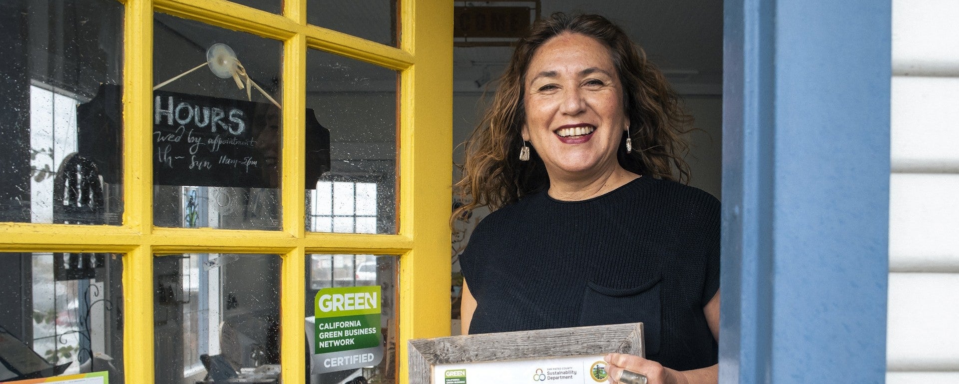 A woman stands at a business entrance holding up a Green Business certificate.