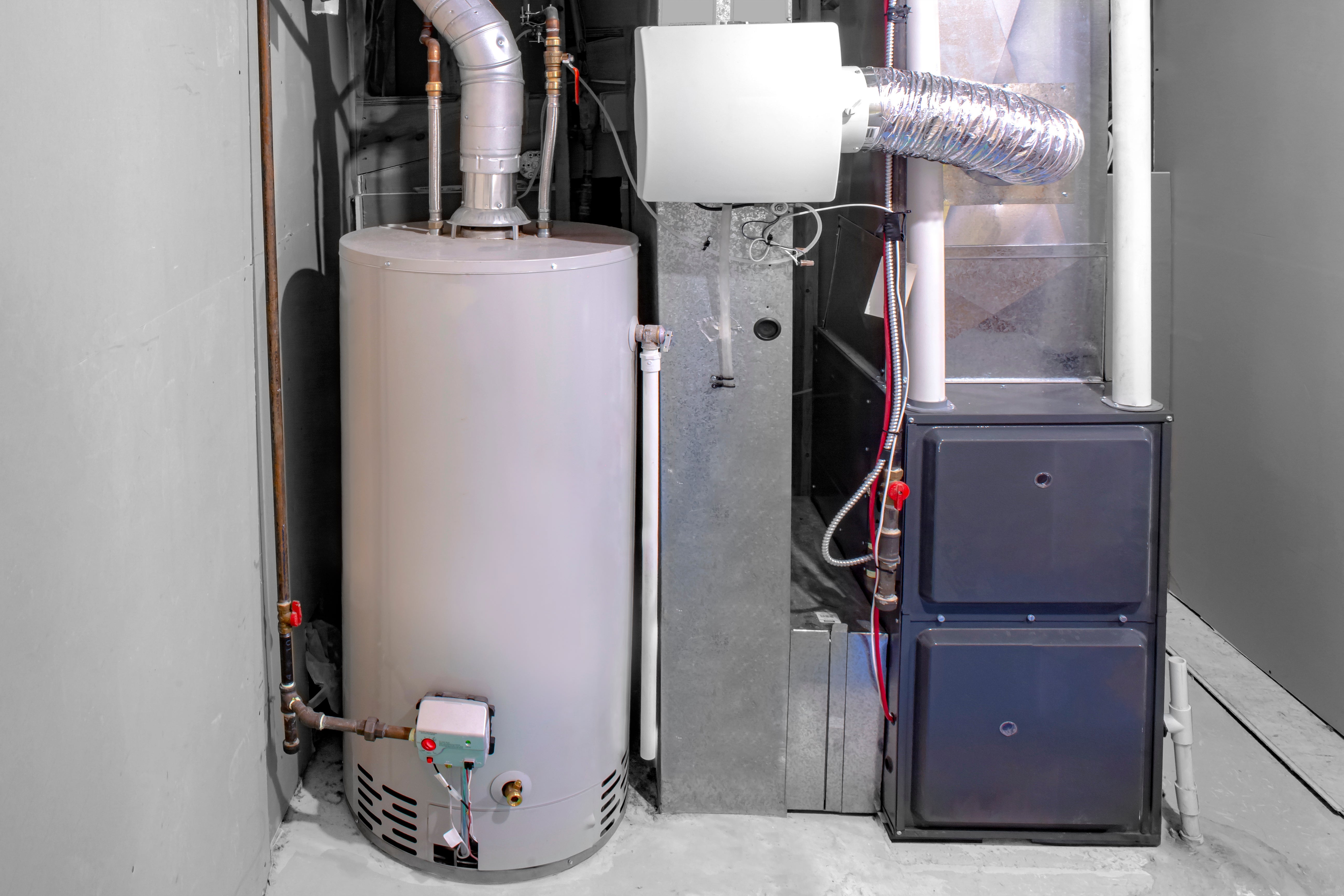 A cylindrical light-gray gas water heater in an enclosed space next to a furnace.