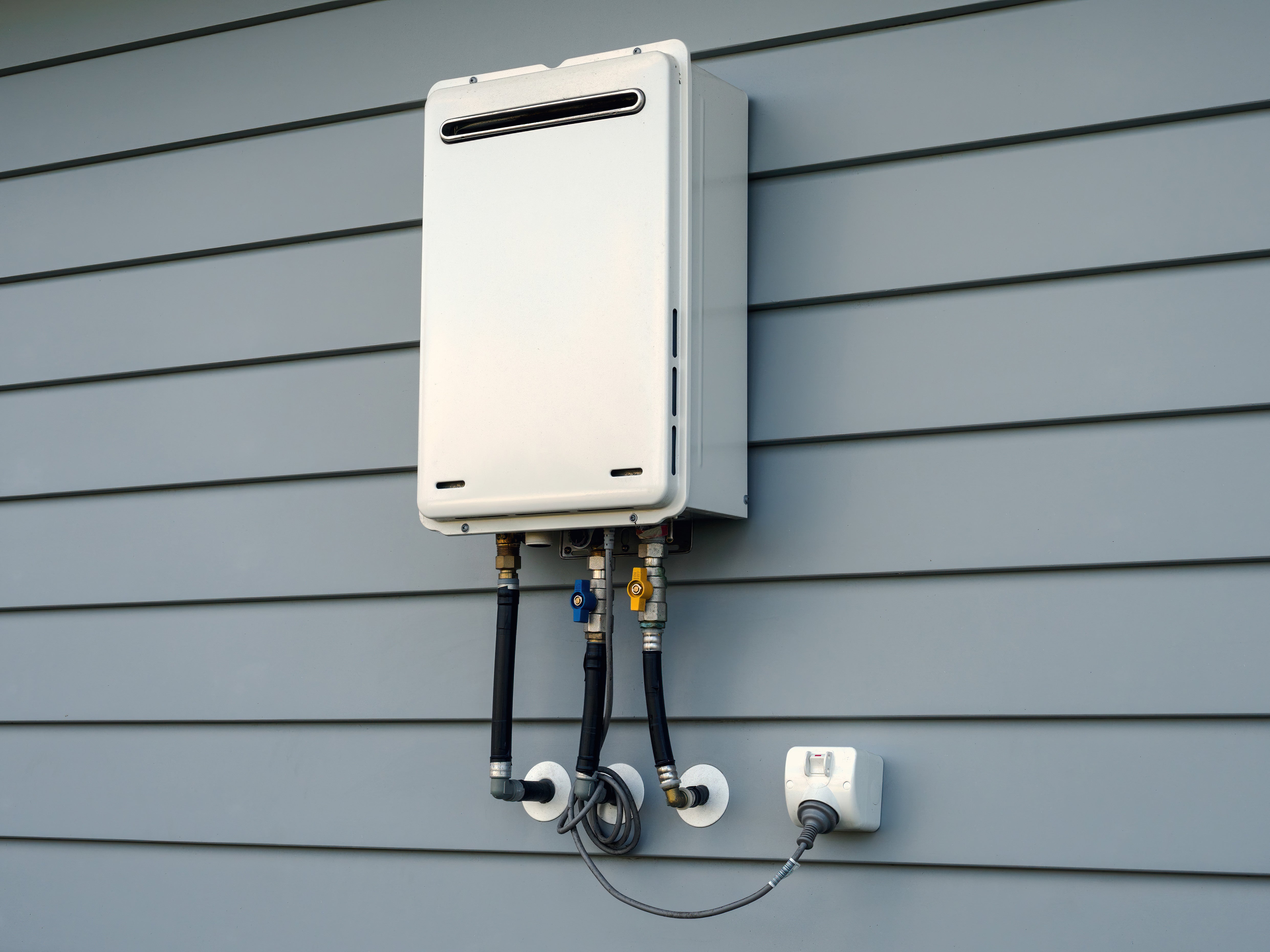 A small white rectangular tankless water heater appliance with pipes coming out of the bottom, mounted on a blue-gray wall.