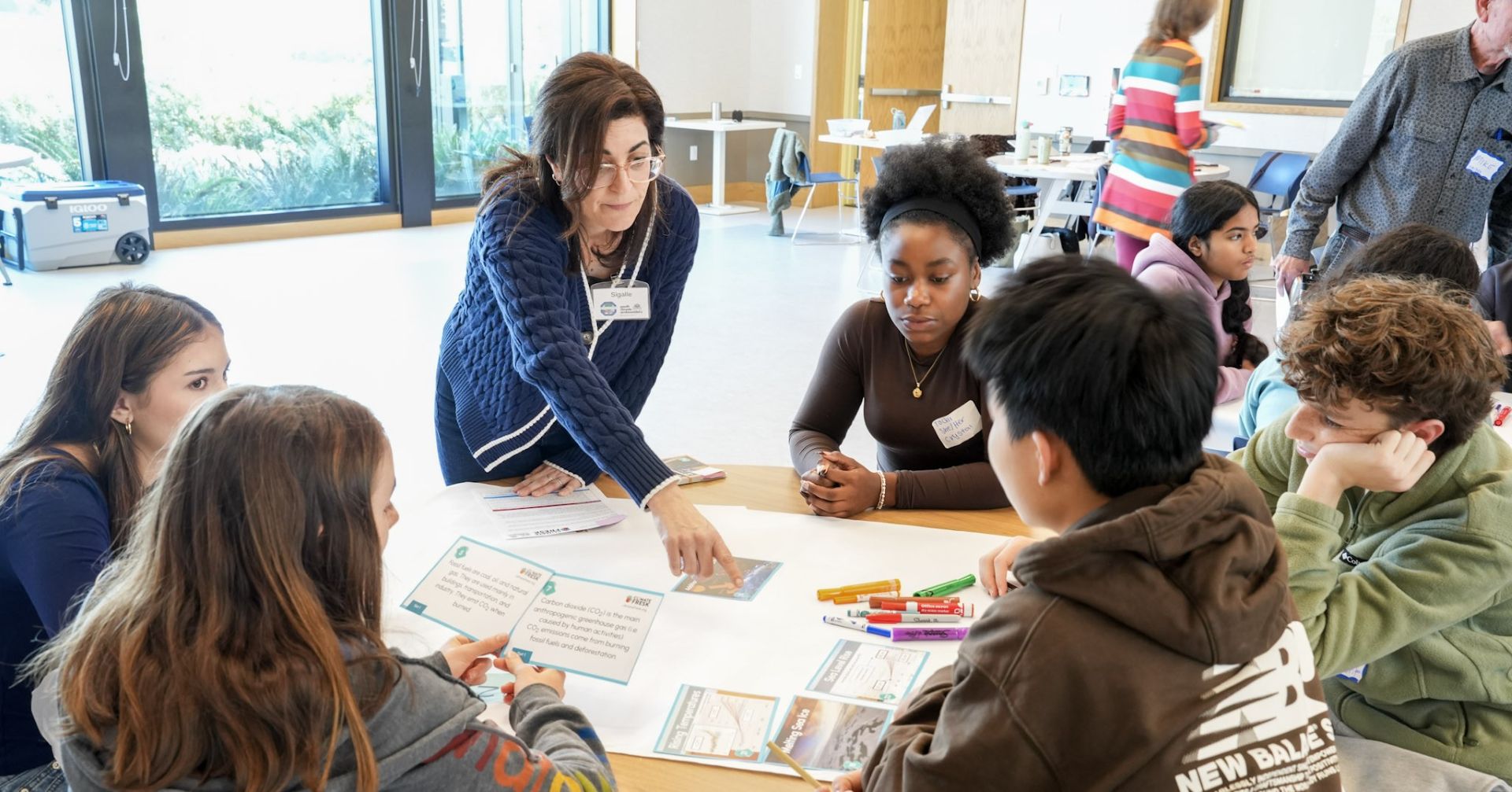 Teacher pointing at an activity on a table with five students looking at her.